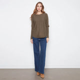 A woman modeling a loose-fitting, long-sleeve olive green sweater paired with dark blue wide-leg jeans and tan pointed-toe flats, standing against a plain white wall.