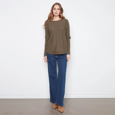 A woman modeling a loose-fitting, long-sleeve olive green sweater paired with dark blue wide-leg jeans and tan pointed-toe flats, standing against a plain white wall.