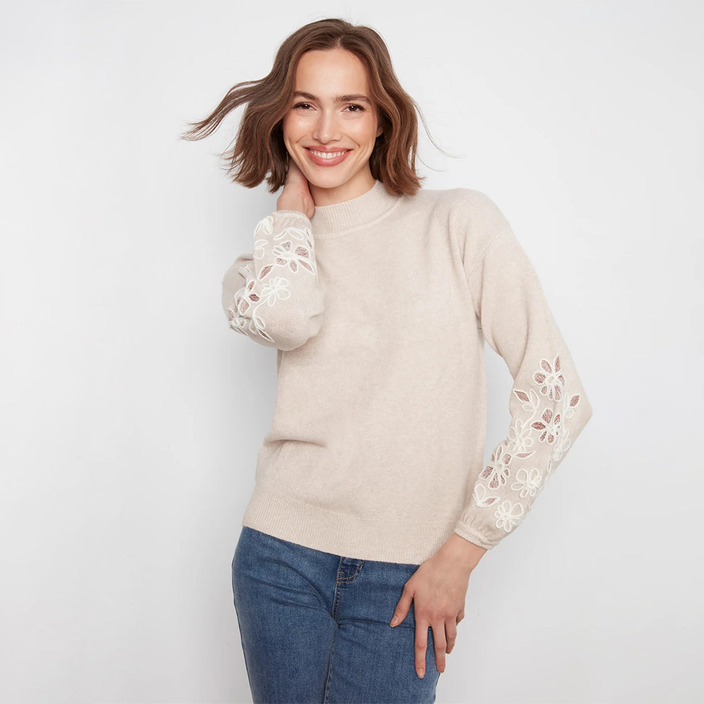 A woman wearing a light beige mock-neck knit sweater with white floral lace embroidery on the sleeves, paired with blue jeans, against a plain white background.