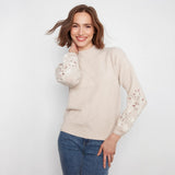 A woman wearing a light beige mock-neck knit sweater with white floral lace embroidery on the sleeves, paired with blue jeans, against a plain white background.