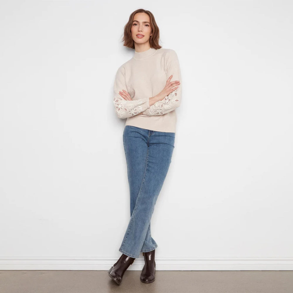 A woman stands against a white wall wearing a cream mock-neck knit sweater with intricate lace sleeves, blue jeans, and dark brown ankle boots.
