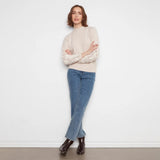A woman stands against a white wall wearing a cream mock-neck knit sweater with intricate lace sleeves, blue jeans, and dark brown ankle boots.