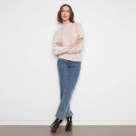 A woman stands against a white wall wearing a cream mock-neck knit sweater with intricate lace sleeves, blue jeans, and dark brown ankle boots.