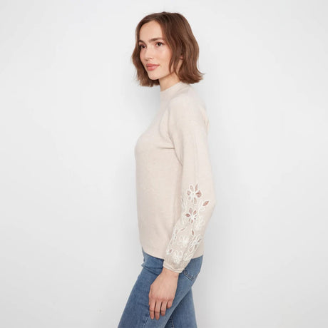 A woman wearing a light beige mock-neck knit sweater with intricate floral lace embroidery on the sleeves, paired with blue jeans, photographed against a clean white studio background.