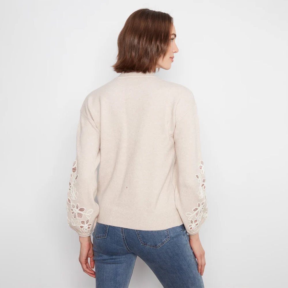 A woman viewed from the back wearing a light beige knit sweater with intricate floral lace cutout detailing on the sleeves, paired with blue jeans, against a plain white studio background.