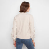 A woman viewed from the back wearing a light beige knit sweater with intricate floral lace cutout detailing on the sleeves, paired with blue jeans, against a plain white studio background.