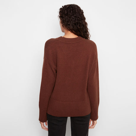 A woman viewed from behind wearing a rust-brown ribbed knit sweater and black pants against a plain white studio background.