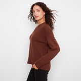 A woman wearing a brown ribbed knit sweater and black pants, photographed from the side against a plain light gray studio background.