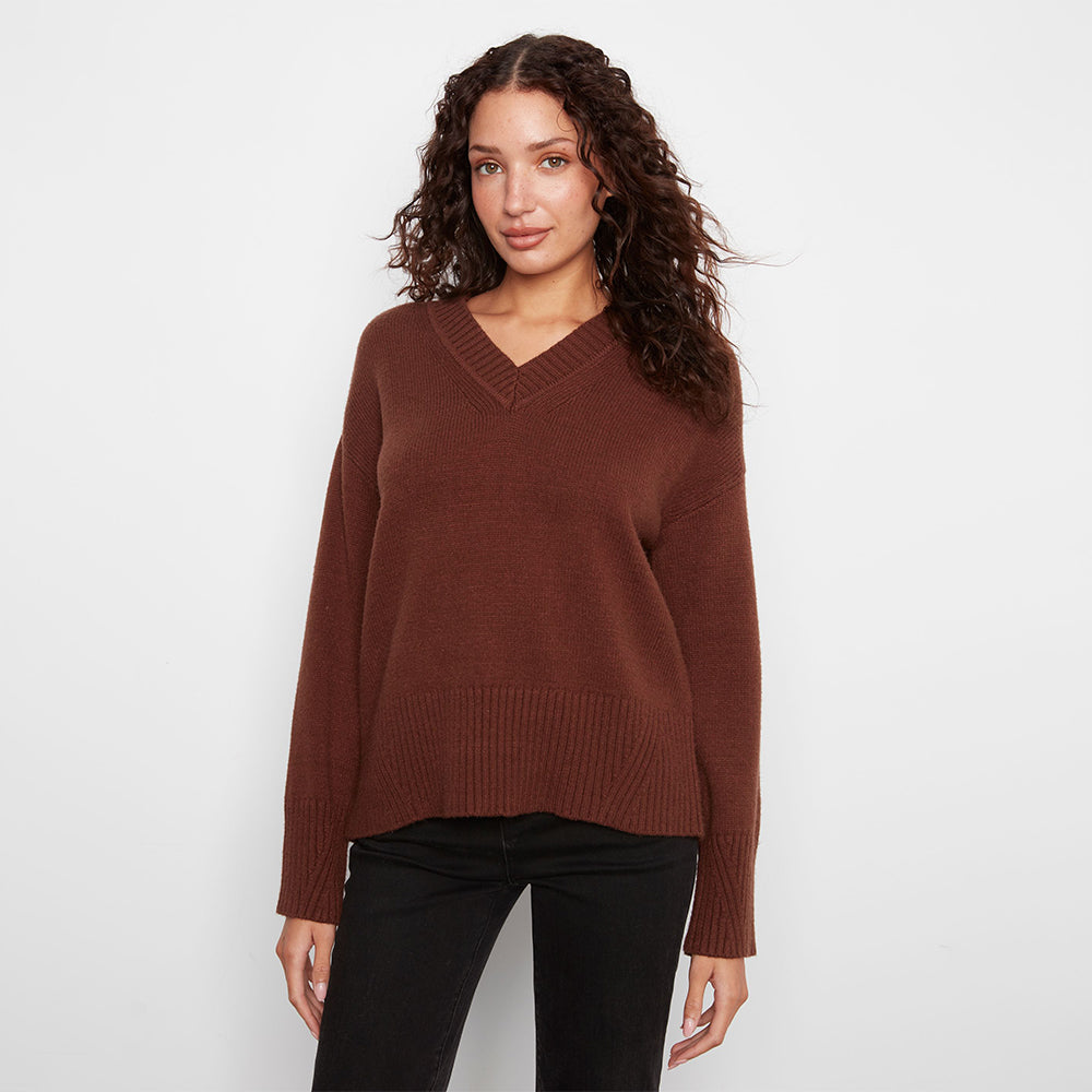 A woman wearing a brown v-neck knit sweater and black pants, standing against a plain light gray studio background.