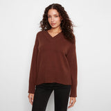 A woman wearing a brown v-neck knit sweater and black pants, standing against a plain light gray studio background.