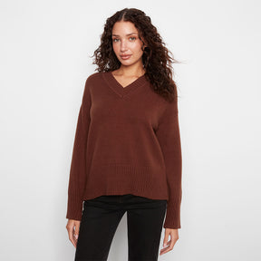 A woman wearing a brown v-neck knit sweater and black pants, standing against a plain light gray studio background.