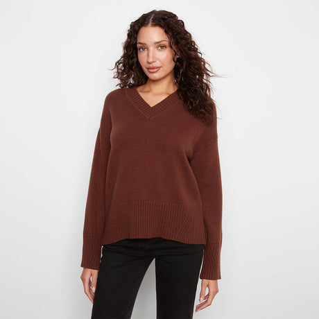 A woman wearing a brown v-neck knit sweater and black pants, standing against a plain light gray studio background.