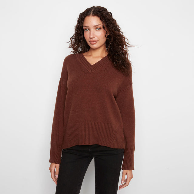 A woman wearing a brown v-neck knit sweater and black pants, standing against a plain light gray studio background.