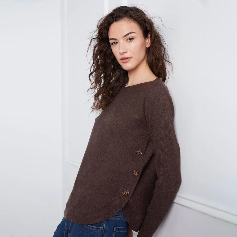A woman wearing a dark brown knit sweater with decorative buttons on the side, paired with blue jeans, standing against a white wall.
