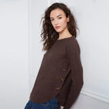 A woman wearing a dark brown knit sweater with decorative buttons on the side, paired with blue jeans, standing against a white wall.