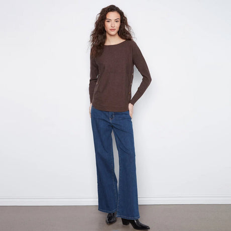 A woman modeling a brown long-sleeve knit top with side pockets and blue wide-leg jeans, standing against a plain white wall.