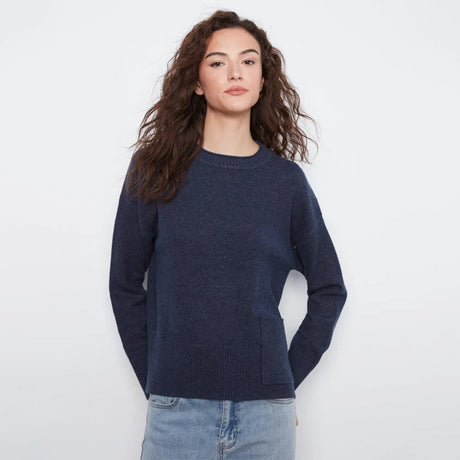 A woman with long, wavy brown hair wearing a dark navy blue crewneck sweater and light blue jeans, standing against a plain white studio background.