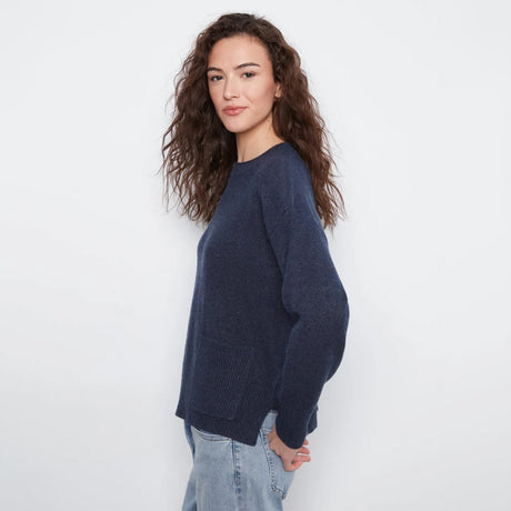 A woman with long, wavy brown hair is shown from a side-back angle wearing a dark navy blue knit sweater and light-wash blue jeans against a plain white studio background.