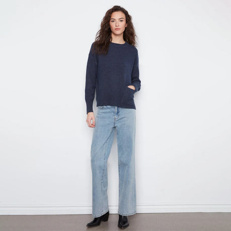 A woman stands against a plain white wall wearing a dark navy long-sleeve knit sweater, light-wash blue straight-leg jeans, and black ankle boots.