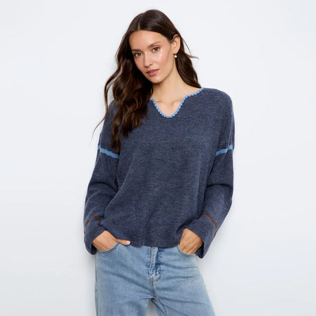 A woman modeling a navy blue knit sweater with light blue trim details on the v-neck and sleeves, paired with light-wash blue jeans against a plain white studio background.