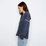 A woman wearing a navy blue knit sweater with light blue and brown stripe accents on the sleeves, paired with light-wash jeans, standing against a plain white wall.