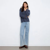 A woman stands against a plain white wall wearing a dark blue-gray knit sweater with light blue wide-leg jeans and brown leather loafers.