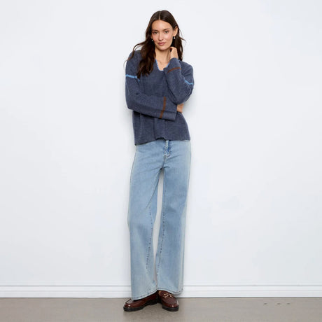 A woman stands against a plain white wall wearing a dark blue-gray knit sweater with light blue wide-leg jeans and brown leather loafers.