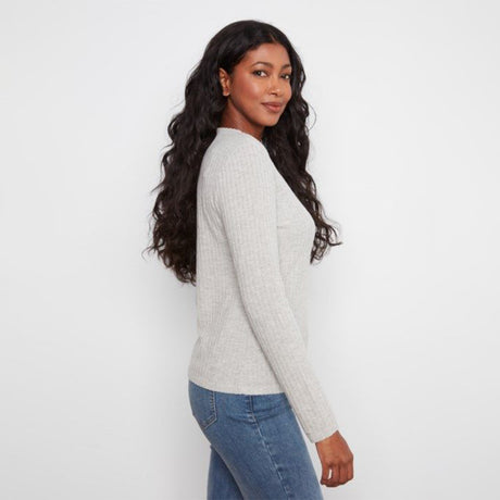 A woman with long, wavy dark hair is shown from a side-back angle wearing a light gray ribbed knit sweater and blue jeans against a plain white studio background.