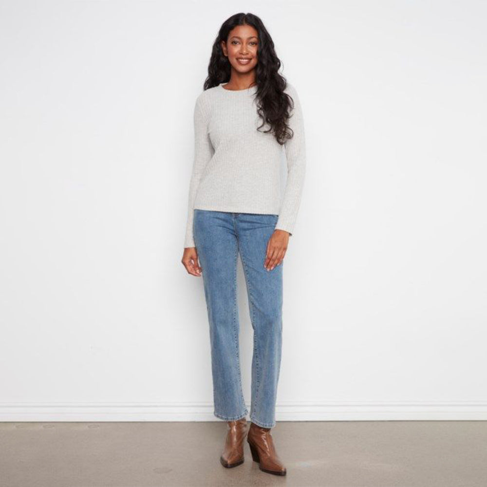 A woman stands against a plain white wall wearing a light heather gray long-sleeve knit sweater, blue straight-leg jeans, and brown ankle boots.