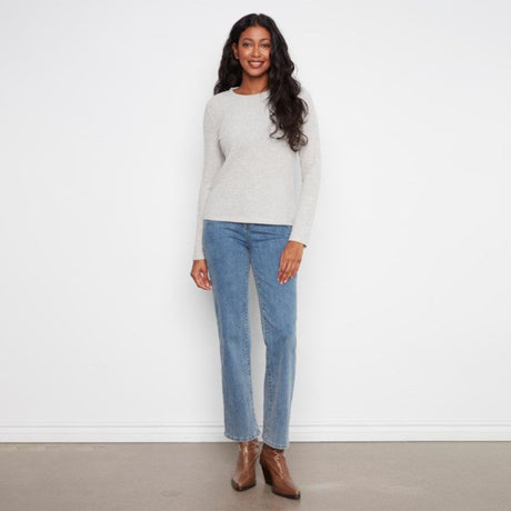 A woman stands against a plain white wall wearing a light heather gray long-sleeve knit sweater, blue straight-leg jeans, and brown ankle boots.