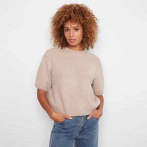 A woman with curly light brown hair wearing a short-sleeve, crew-neck knit sweater in a light beige color, paired with blue jeans, standing against a plain white background.