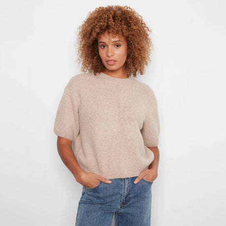 A woman with curly light brown hair wearing a short-sleeve, crew-neck knit sweater in a light beige color, paired with blue jeans, standing against a plain white background.