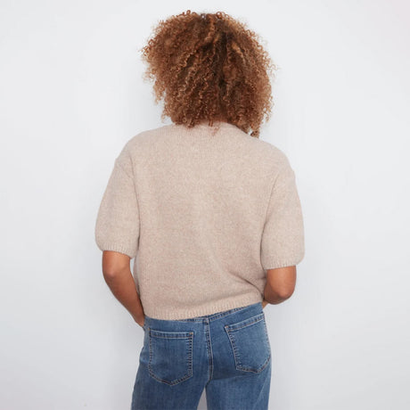 A person viewed from behind wearing a short-sleeve knit sweater in a light beige color and blue jeans, standing against a plain white wall.