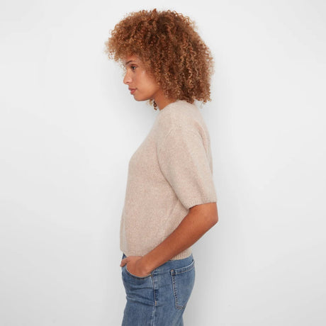 A woman with curly brown hair is shown in profile wearing a short-sleeve knit sweater in a light beige color and blue jeans, standing against a plain white background.