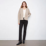 A woman stands against a white wall wearing a light beige knit cardigan over a white top, black straight-leg jeans, and black ankle boots.
