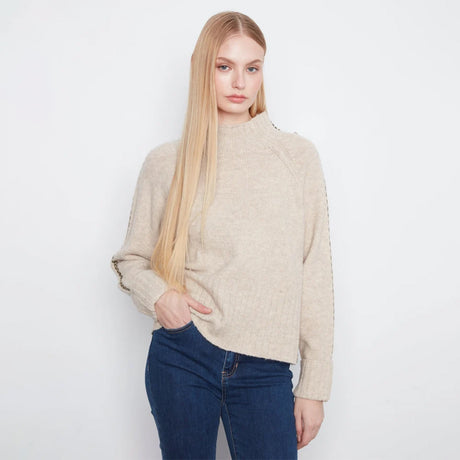A blonde model wearing a beige mock neck knit sweater with gold chain detailing along the shoulders and sleeves, paired with dark blue jeans, against a plain white background.