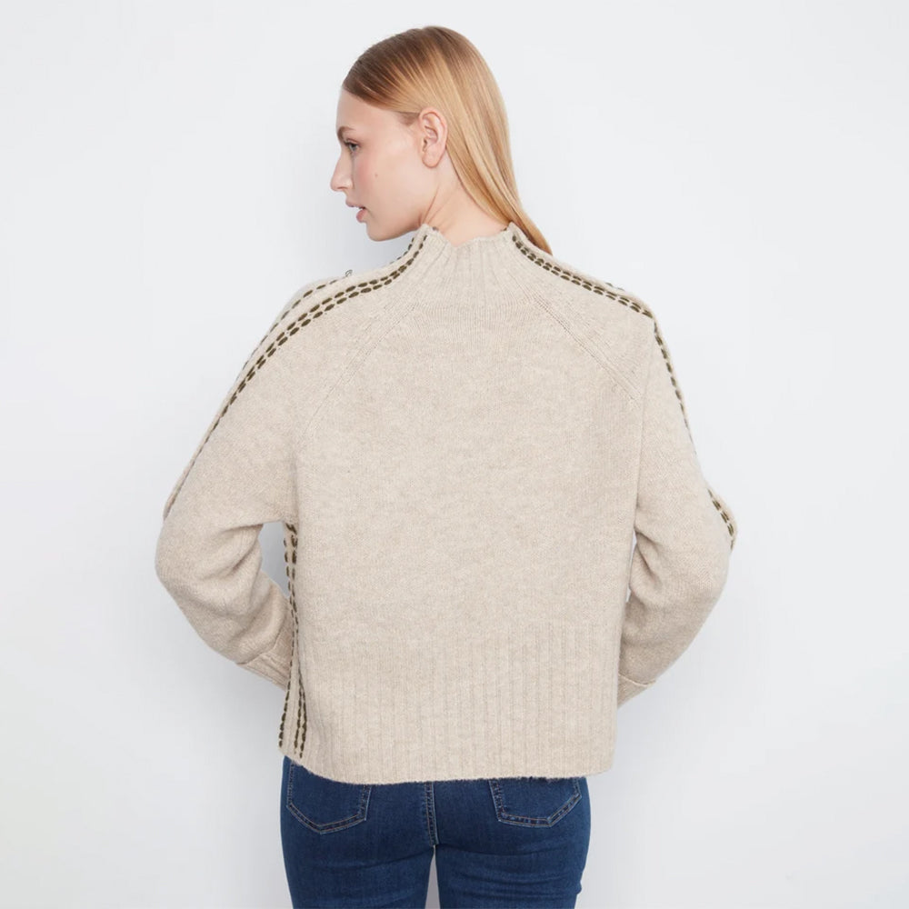 A woman shown from the back wearing a light beige knit sweater with decorative chain detailing along the shoulders and sides, paired with blue jeans, against a plain white background.