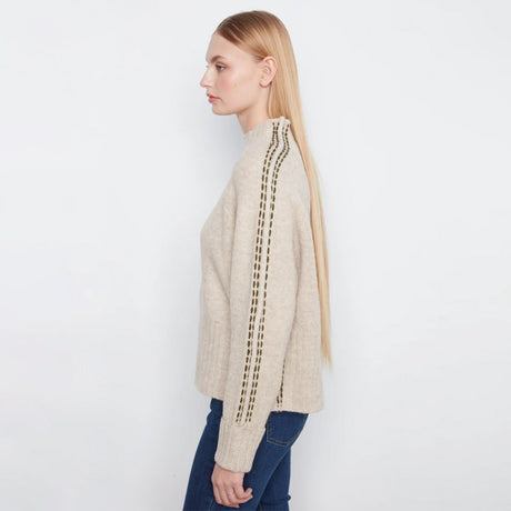 A woman in profile wearing a beige knit sweater with vertical brown textured stripe details on the sleeves, paired with dark blue jeans, against a plain white studio background.