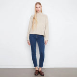 A blonde model stands against a white wall wearing a beige ribbed mock-neck sweater, dark blue skinny jeans, and brown leather loafers.