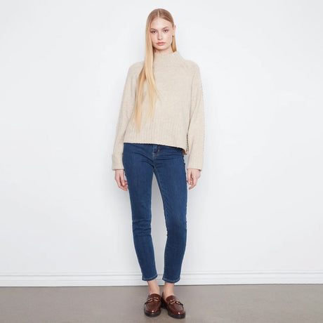 A blonde model stands against a white wall wearing a beige ribbed mock-neck sweater, dark blue skinny jeans, and brown leather loafers.