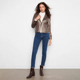 A woman stands against a plain white wall wearing a metallic taupe-brown leather jacket, a white turtleneck, blue skinny jeans, and dark brown ankle boots.