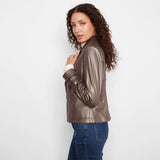 A woman with curly brown hair models a metallic bronze faux-leather jacket, paired with a white collared shirt and blue jeans, against a plain white studio background.