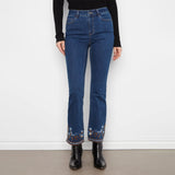 A person wearing dark blue flare jeans with colorful floral embroidery at the hem, paired with a black long-sleeve top and black ankle boots, standing against a white wall.
