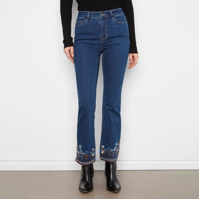 A person wearing dark blue flare jeans with colorful floral embroidery at the hem, paired with a black long-sleeve top and black ankle boots, standing against a white wall.