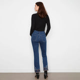 A woman is shown from behind wearing dark blue flare jeans with floral embroidery at the hem, a black ribbed turtleneck sweater, and black ankle boots, standing against a plain white wall.