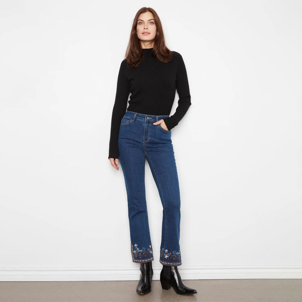 A woman stands against a white wall wearing a black ribbed mock-neck long-sleeve top and blue jeans with floral embroidery at the hem, paired with black ankle boots.