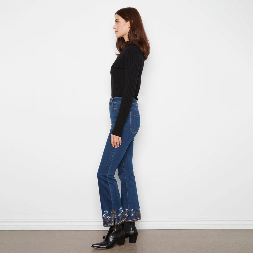 A woman stands in profile wearing a black ribbed long-sleeve top, blue flared jeans with floral embroidery at the hem, and black ankle boots against a plain white wall.