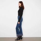 A woman stands in profile wearing a black ribbed long-sleeve top, blue flared jeans with floral embroidery at the hem, and black ankle boots against a plain white wall.
