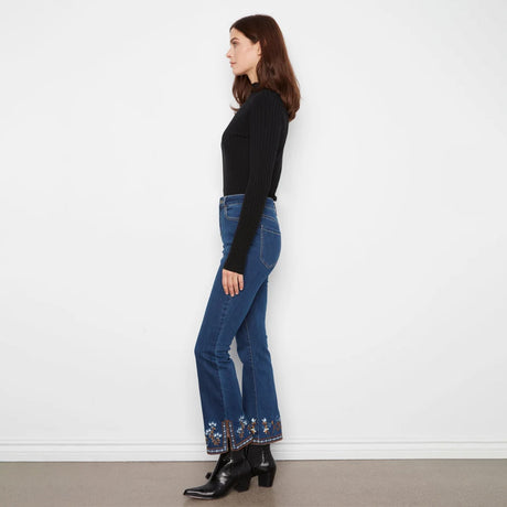 A woman stands in profile wearing a black ribbed long-sleeve top, blue flared jeans with floral embroidery at the hem, and black ankle boots against a plain white wall.