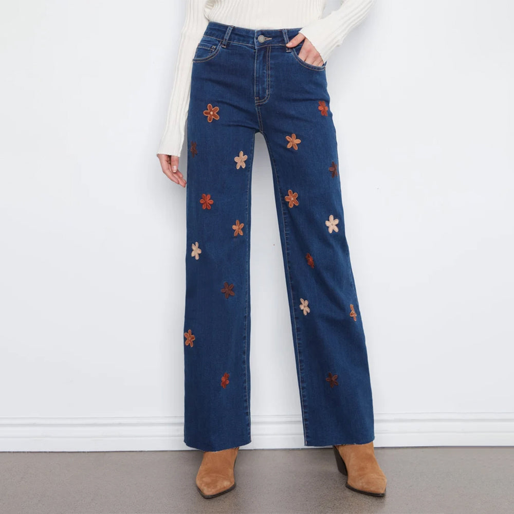 A person wearing dark blue wide-leg jeans with embroidered orange, red, and cream flowers, paired with a white ribbed sweater and tan ankle boots, standing against a white wall.
